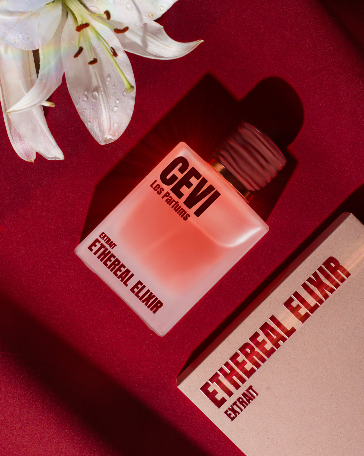 A bottle of CEVI Les Fabriqs Ethereal Elixir perfume with a white cap, displayed next to its white packaging box on a deep red background with a white lily flower in the corner.