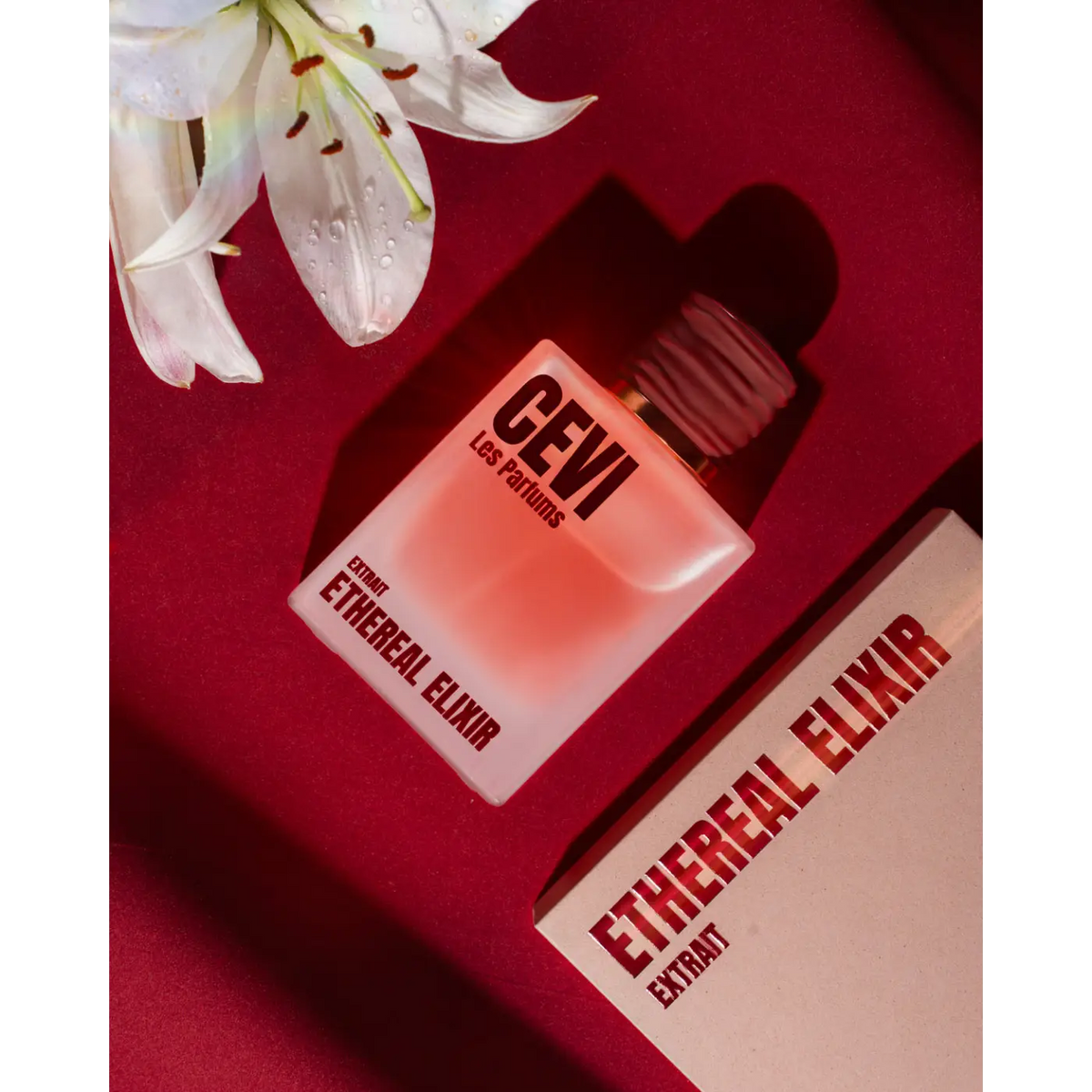 A translucent pink perfume bottle with a gold cap labeled gevi les parfums ethereal elixir beside its box on red surface